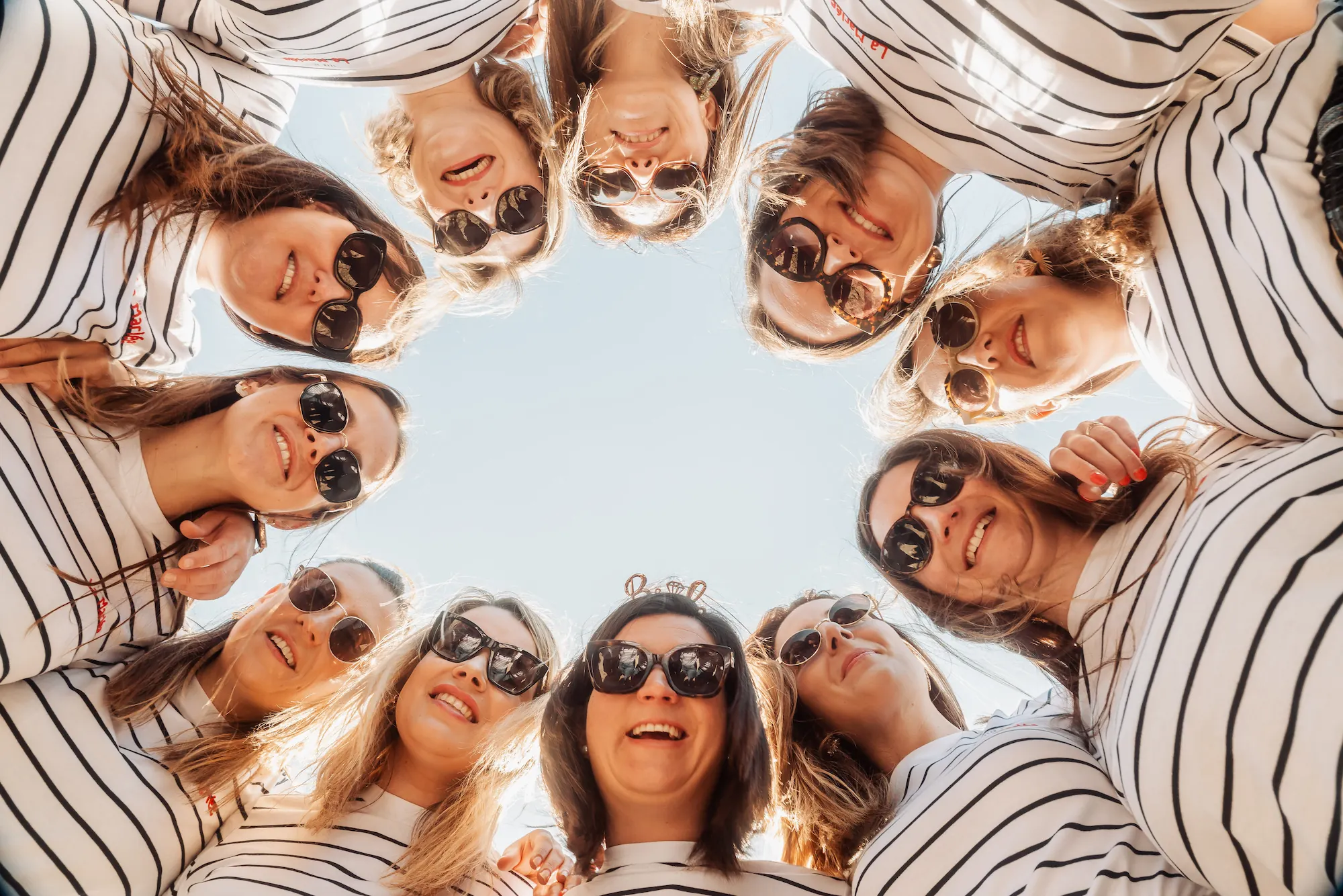 Groupe de femmes en marinière formant un cercle, photographiées du dessous avec le ciel bleu au centre, ambiance solaire
