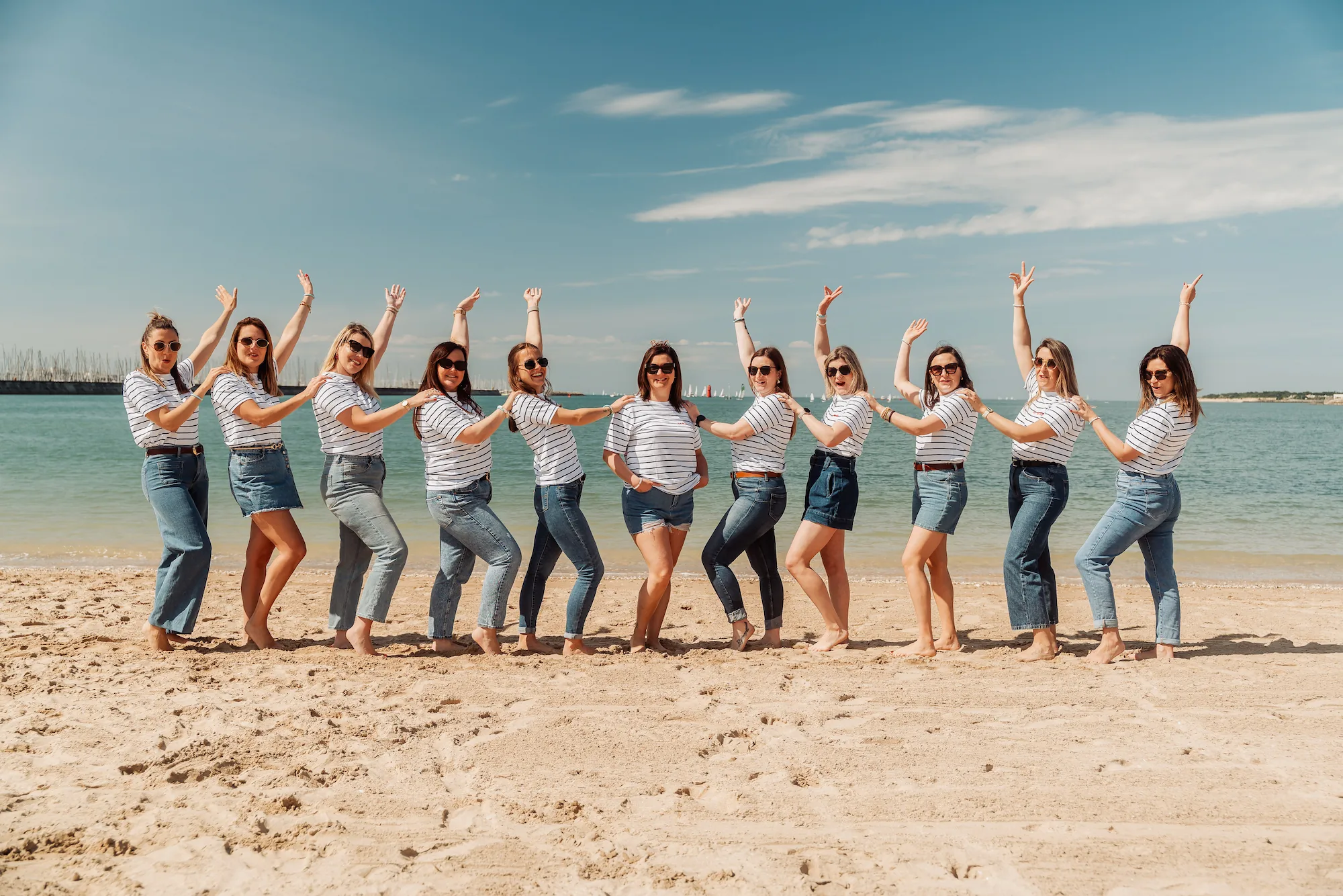 Groupe de femmes en tee-shirt rayé marinière et jean, bras levés, souriantes sur la plage de La Rochelle avec le port en arrière-plan