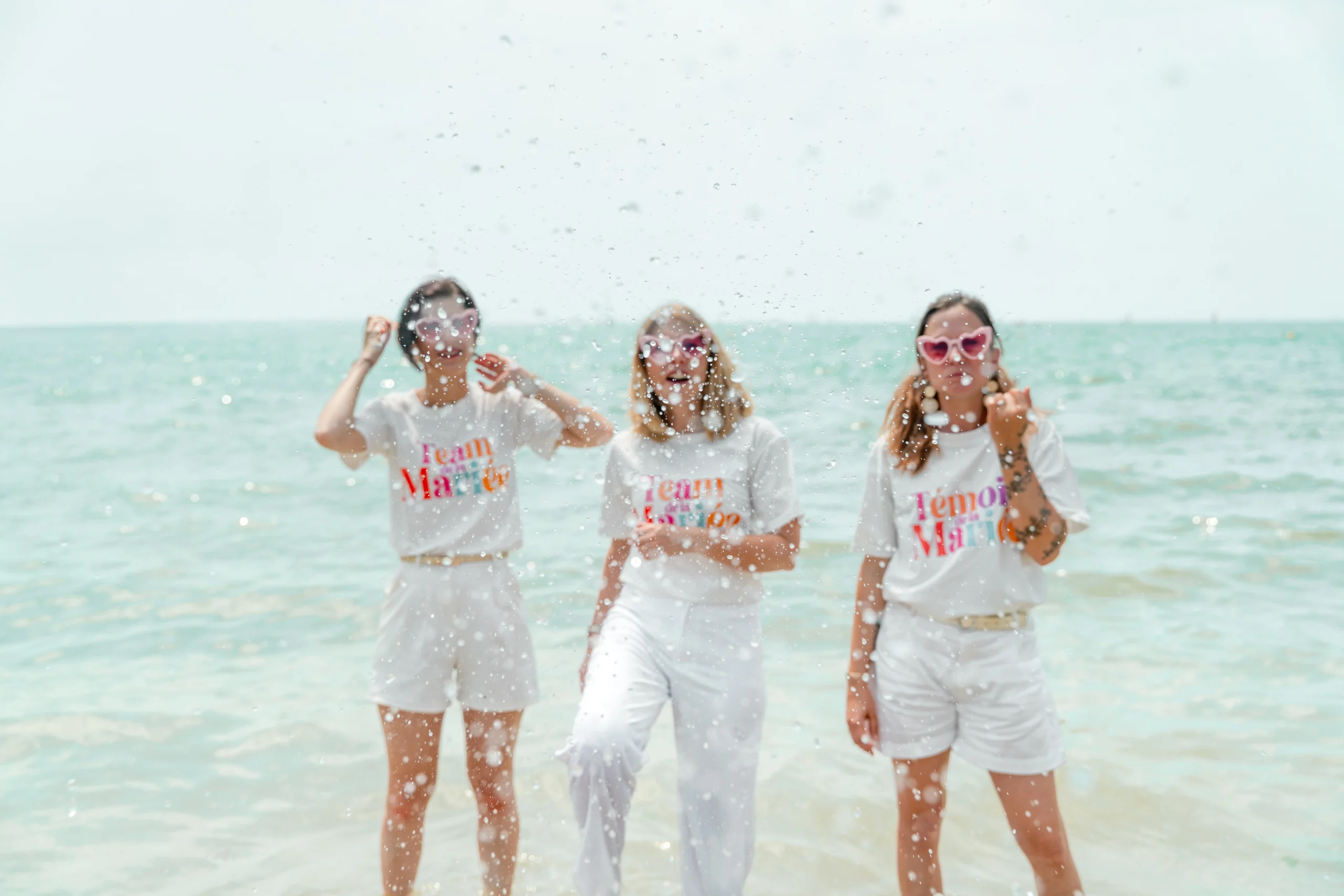 Trois amies en tenue blanche et lunettes cœur s'éclaboussent dans la mer pendant une séance photo EVJF à La Rochelle